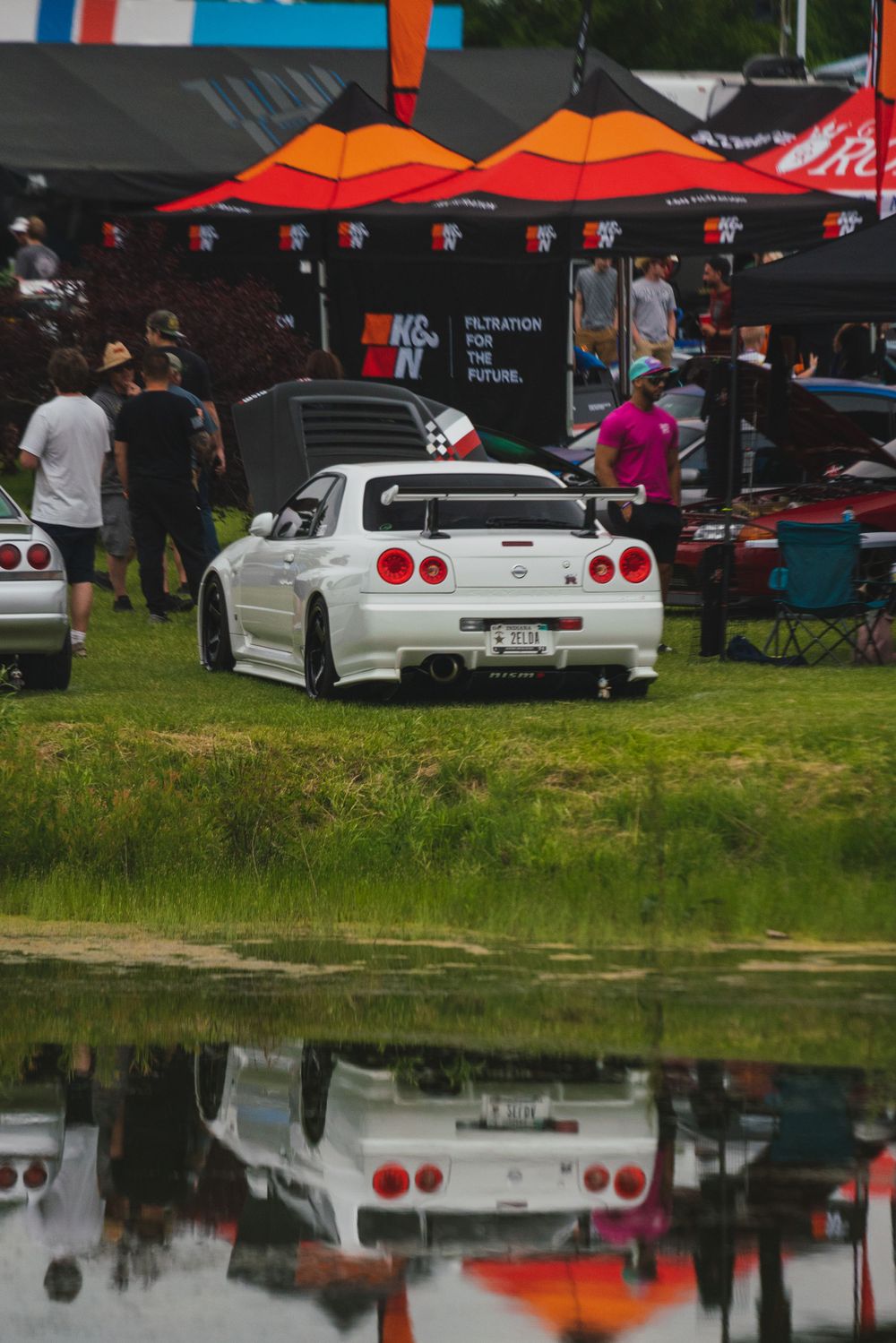 White Nissan Skyline GT-R reflected in puddle at car show with tents in background.