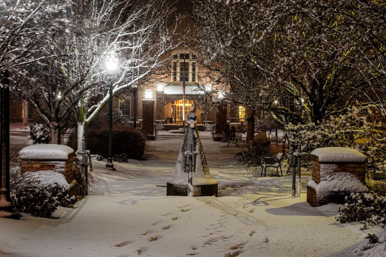 Snow covered walkway leading to an illuminated building entrance at night with trees and lamp posts.