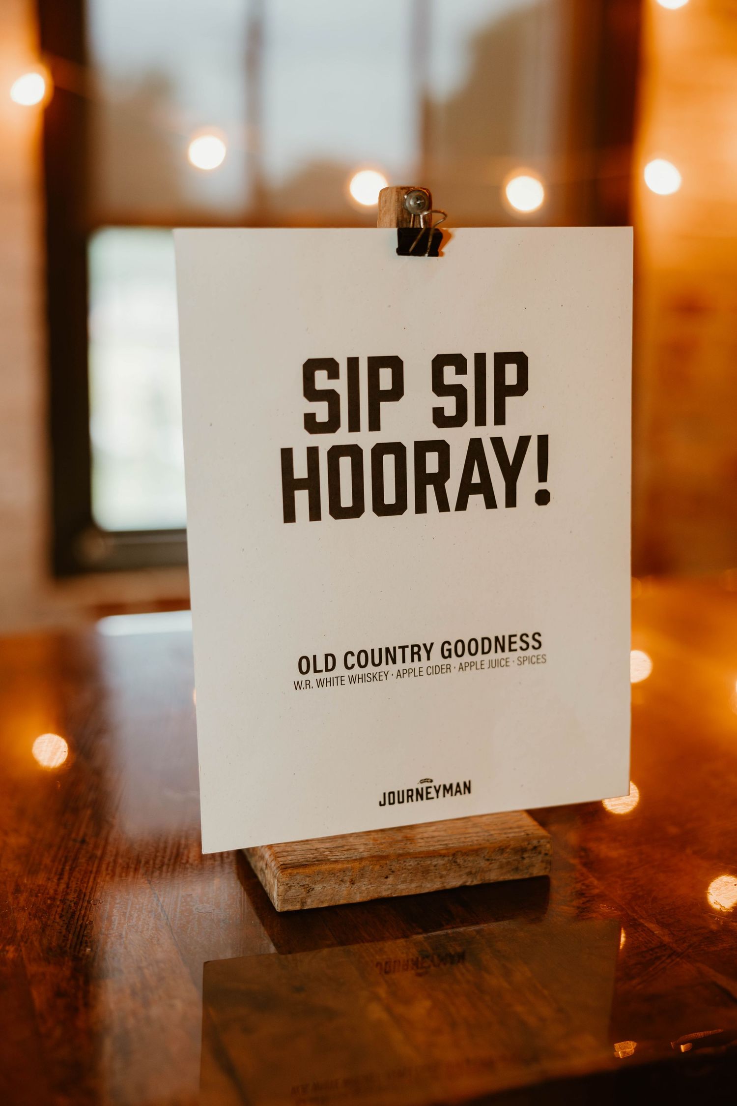 White sign with 'Sip Sip Hooray!' text displayed on wooden stand with warm bokeh lights in background.