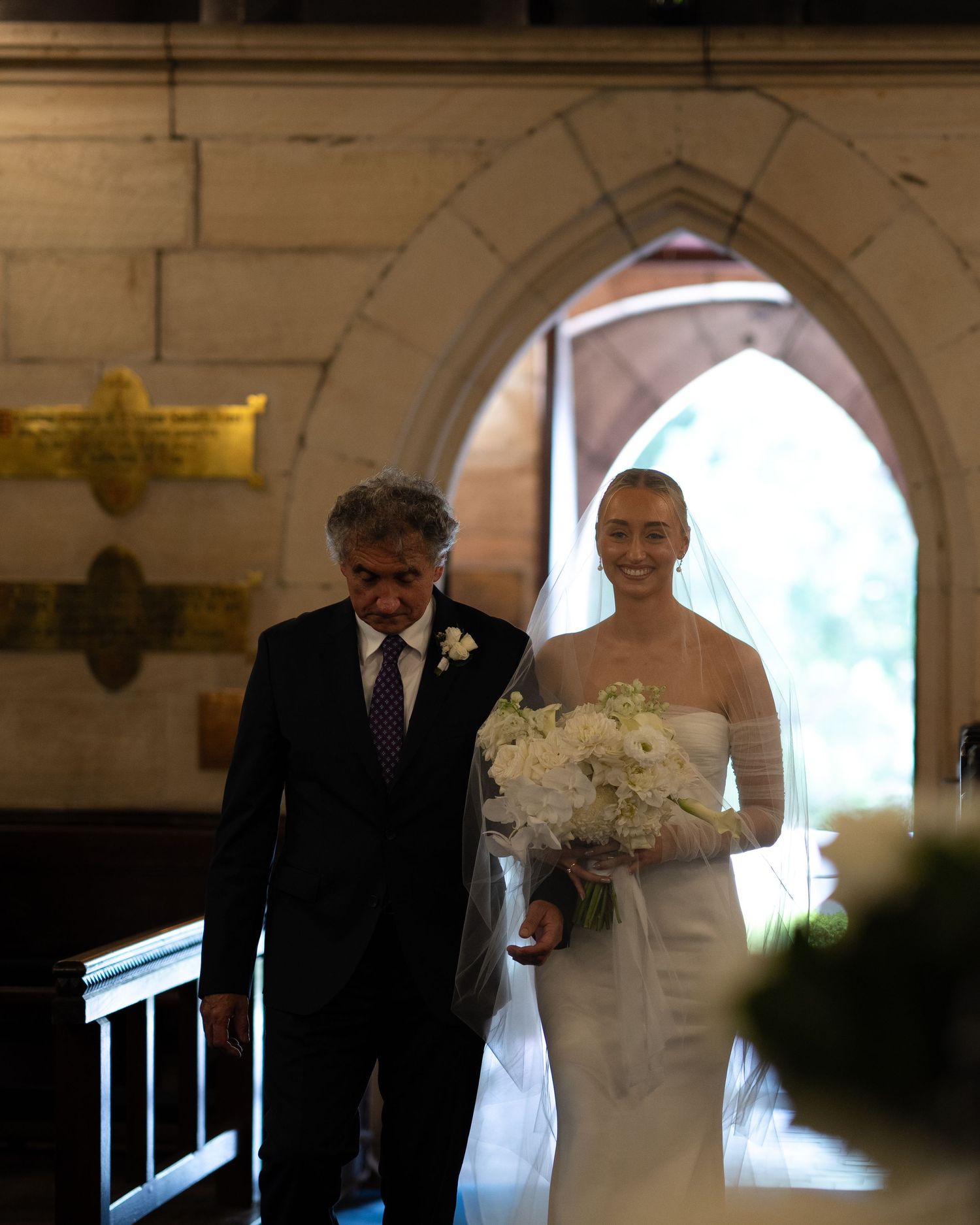 Father walks bride down church aisle holding white bouquet.