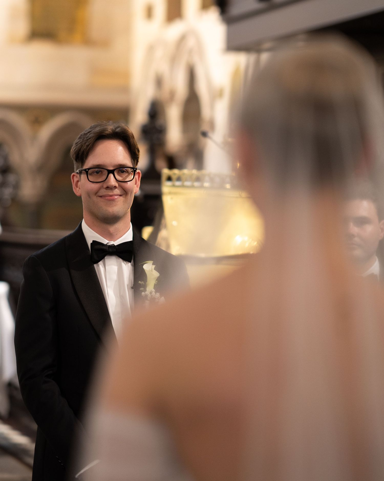 Groom in tuxedo and glasses smiles during wedding ceremony.