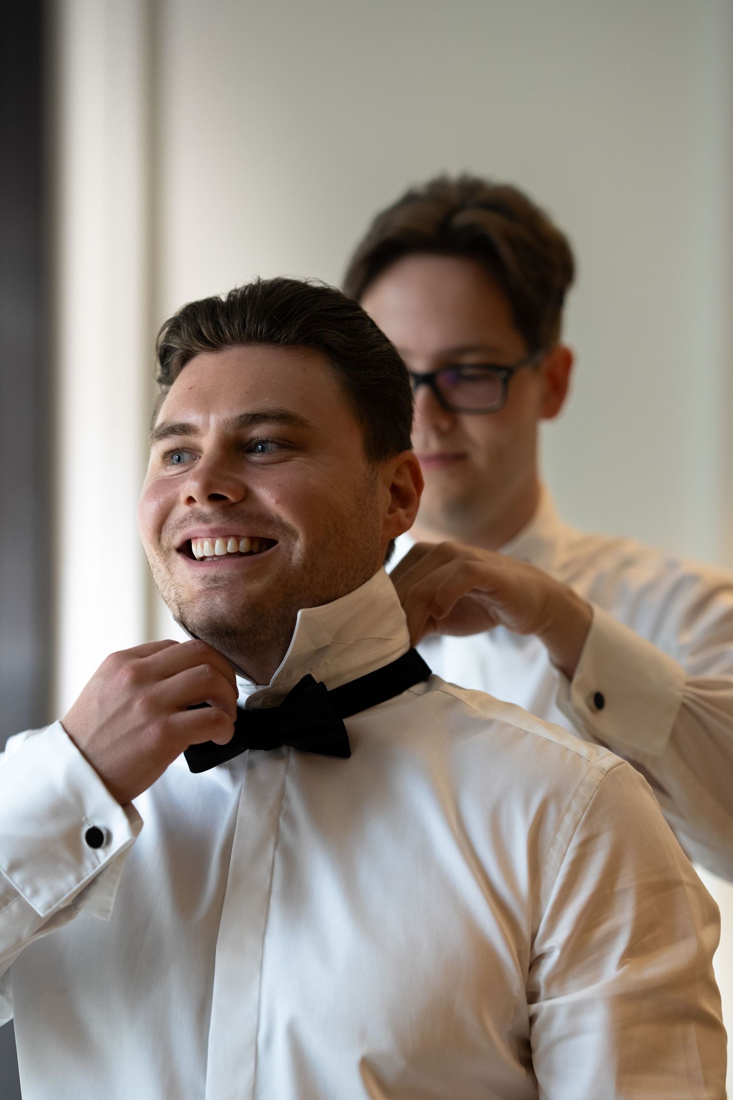 Groom adjusts his bowtie while getting ready with help from his best man.