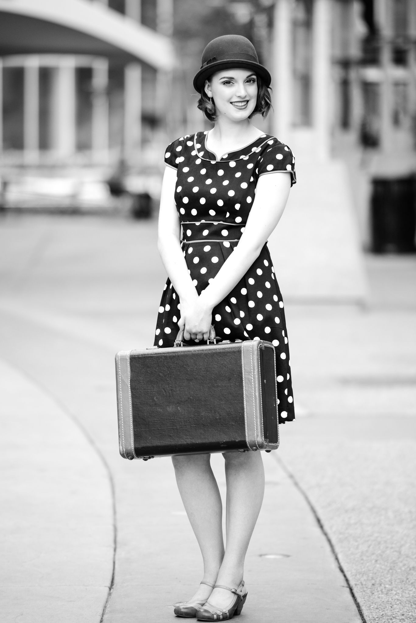 Retro black and white portrait with polka dot dress and vintage suitcase on sidewalk.