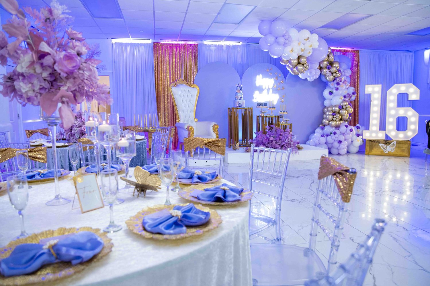 Elegant sweet sixteen party venue decorated in purple and blue lighting with crystal chairs and floral centerpieces.