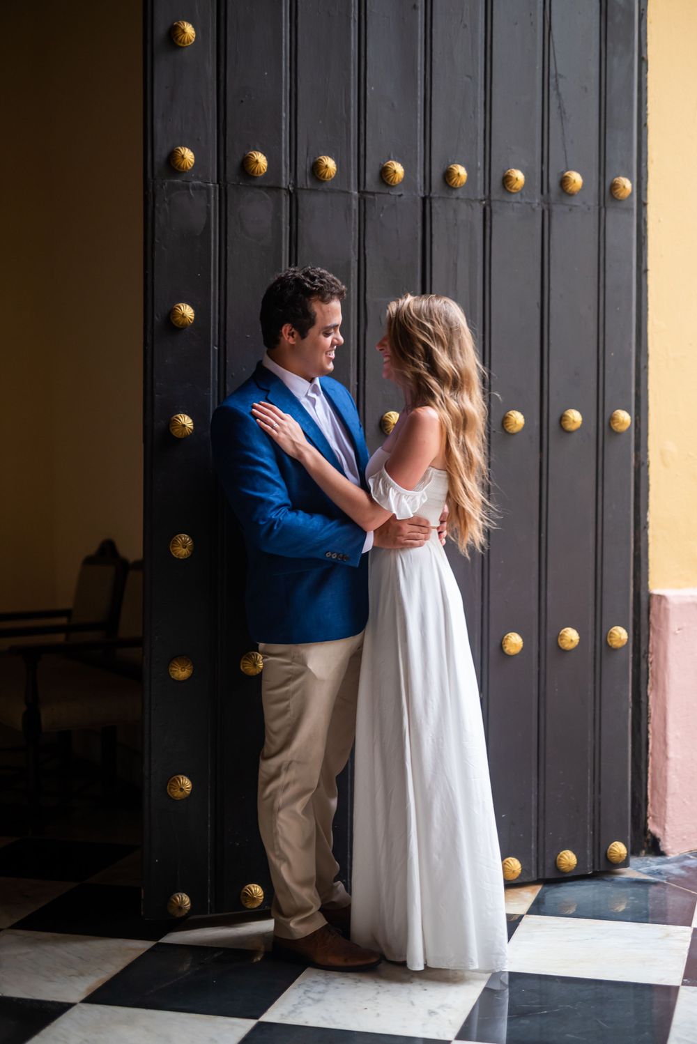 A couple poses against a decorative black door with brass studs in an elegant setting.
