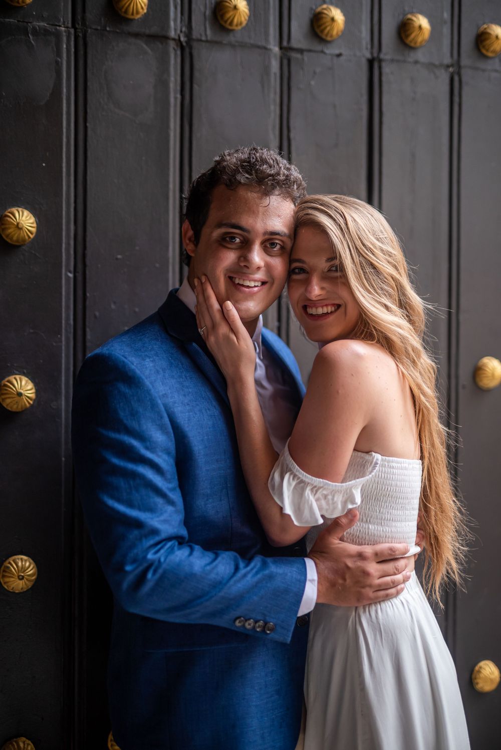 A couple in formal attire embracing in front of a studded black door.