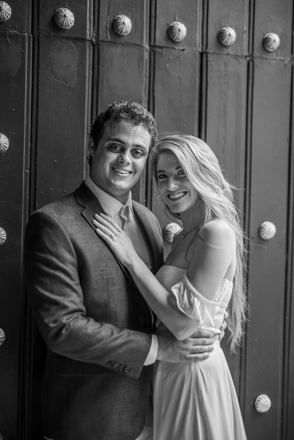 A black and white portrait of a couple posing against a decorative door with metal studs.