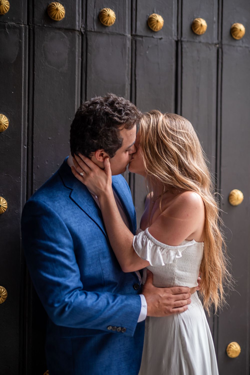 A romantic couple shares a tender kiss against a dark door with gold studs at a beautiful location.