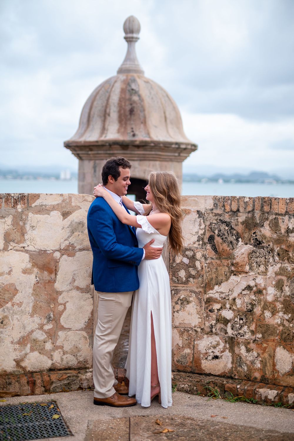 A couple embraces by a historic domed turret overlooking the ocean.