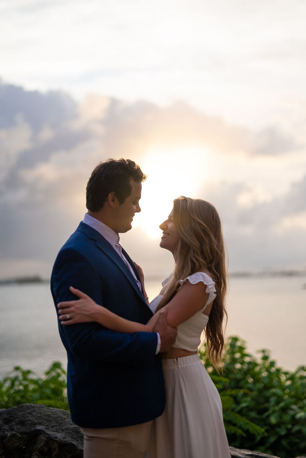 A romantic couple embraces at sunset by the ocean with soft clouds in the background.