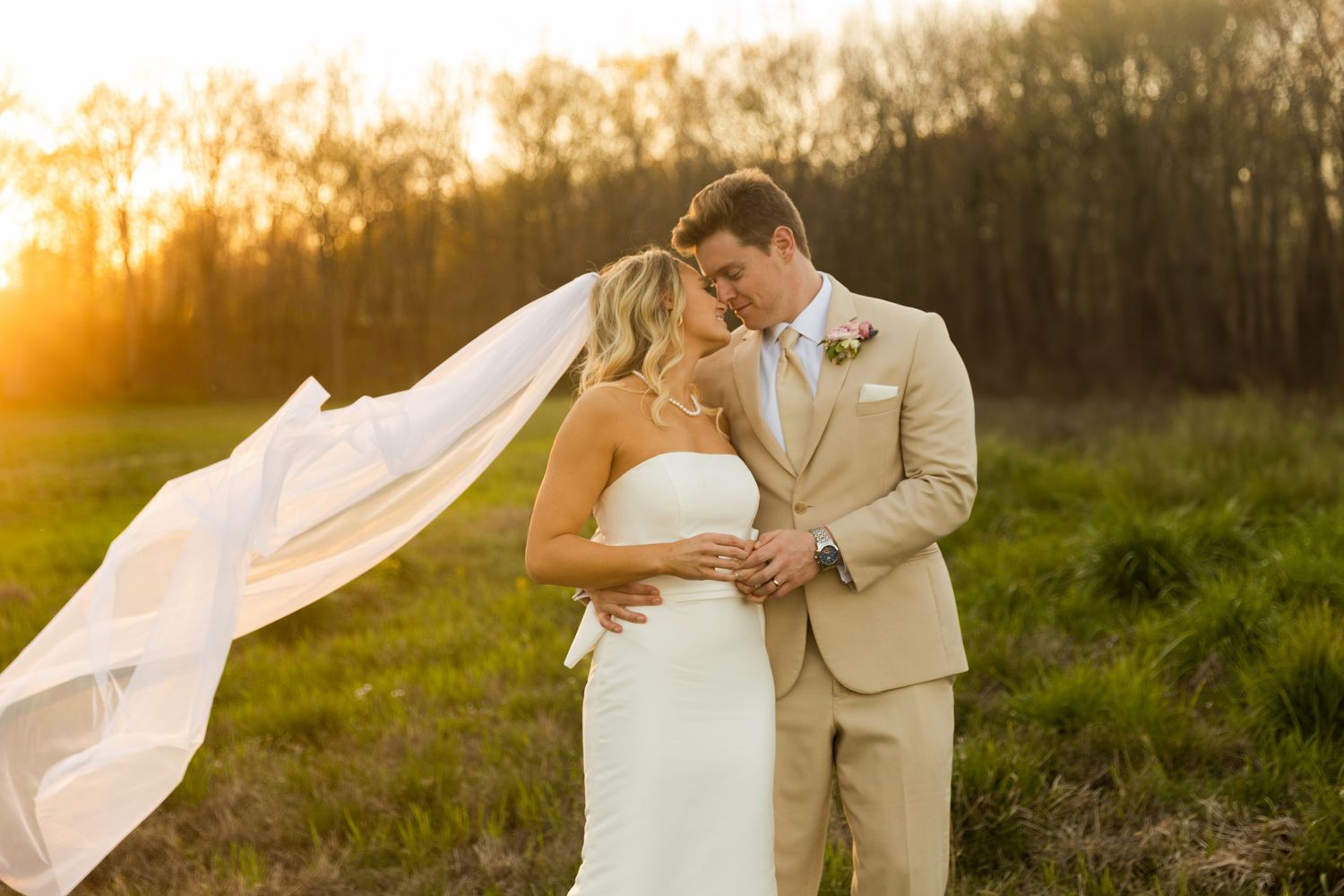 Romantic sunset wedding portrait with flowing veil in rustic outdoor setting.