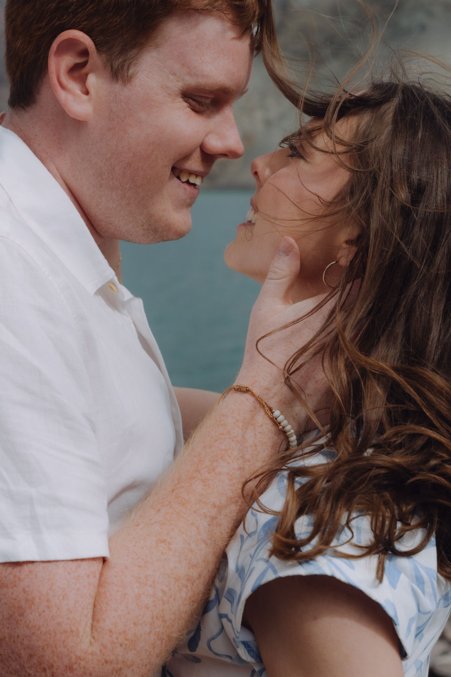Happy couple smiling and embracing closely outdoors near water with wind blowing through hair.