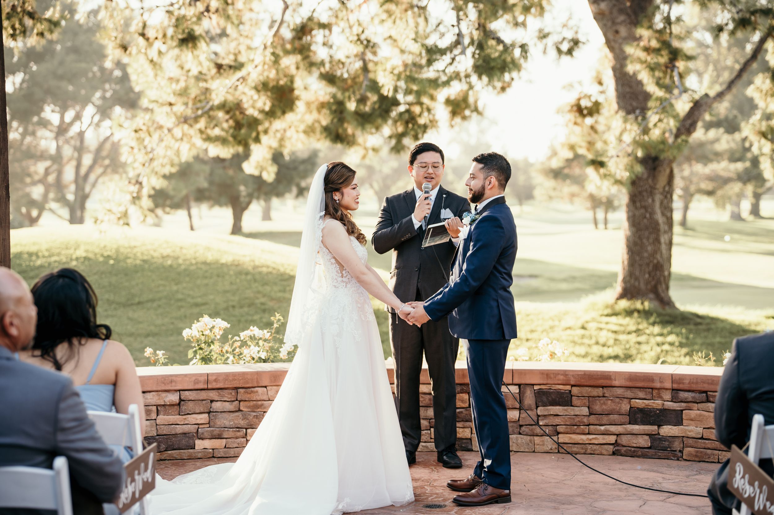 Outdoor wedding ceremony under trees with guests seated on white chairs watching the proceedings.