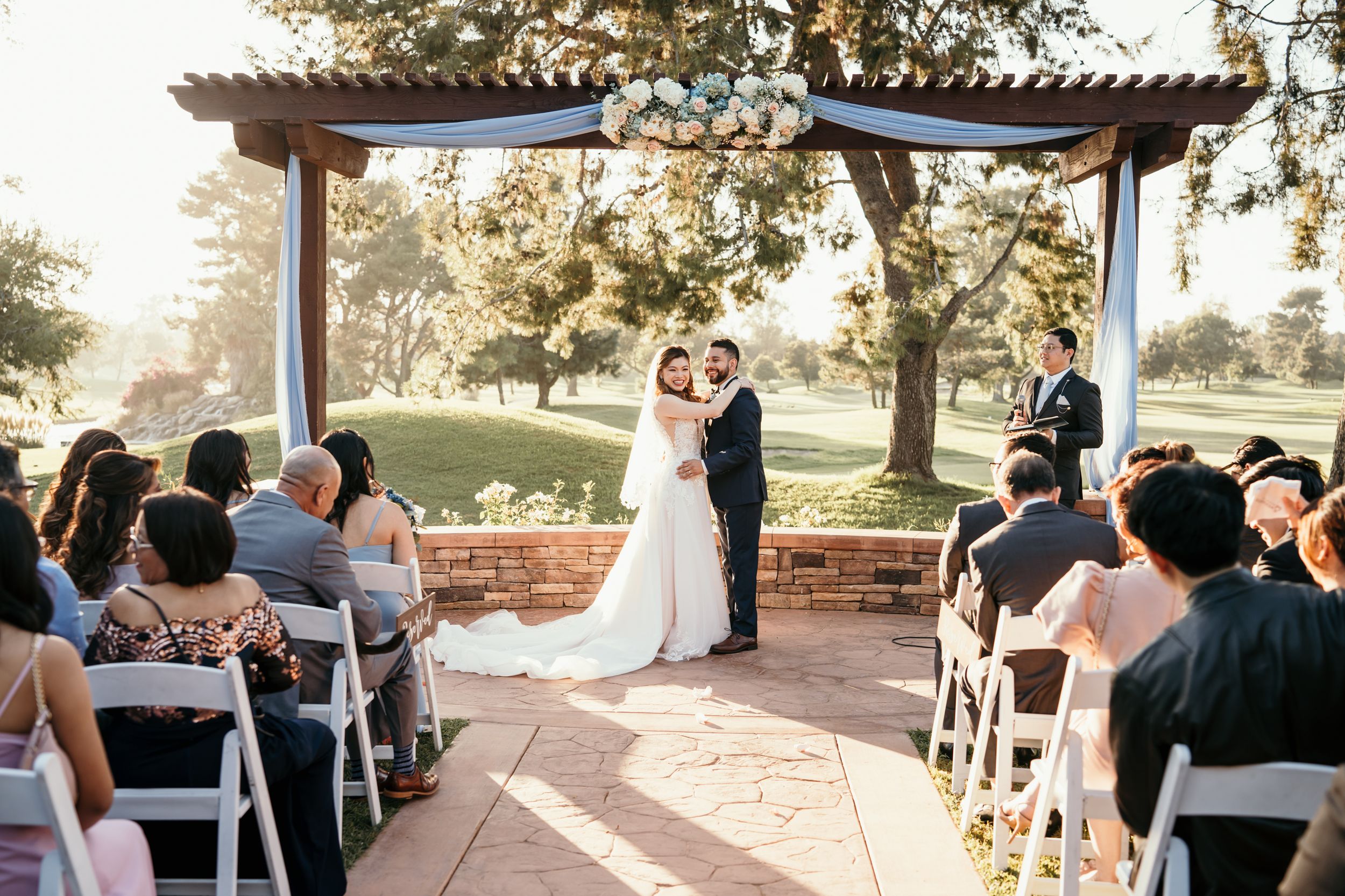 Wide shot of outdoor wedding ceremony with guests seated under a wooden pergola.