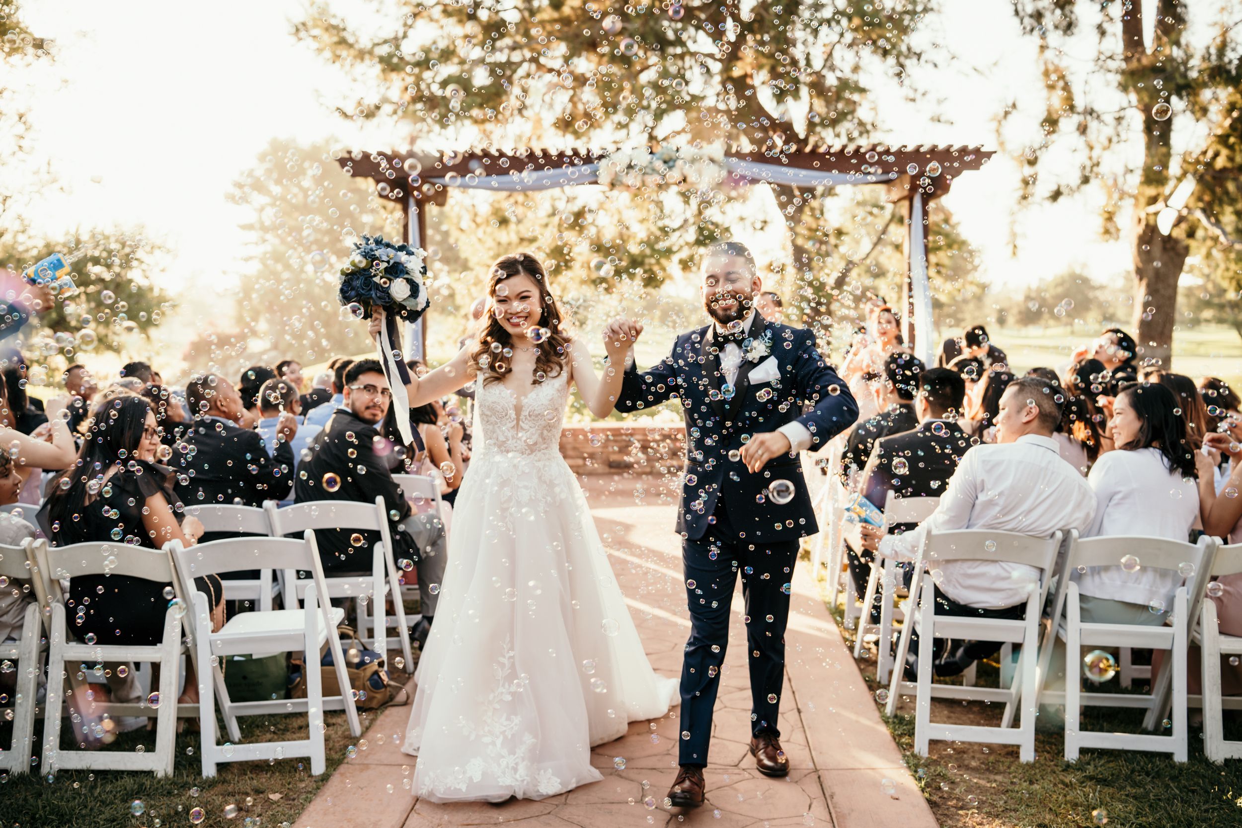 Newlyweds joyfully walk down the aisle at outdoor wedding ceremony with white chairs and floral arch.