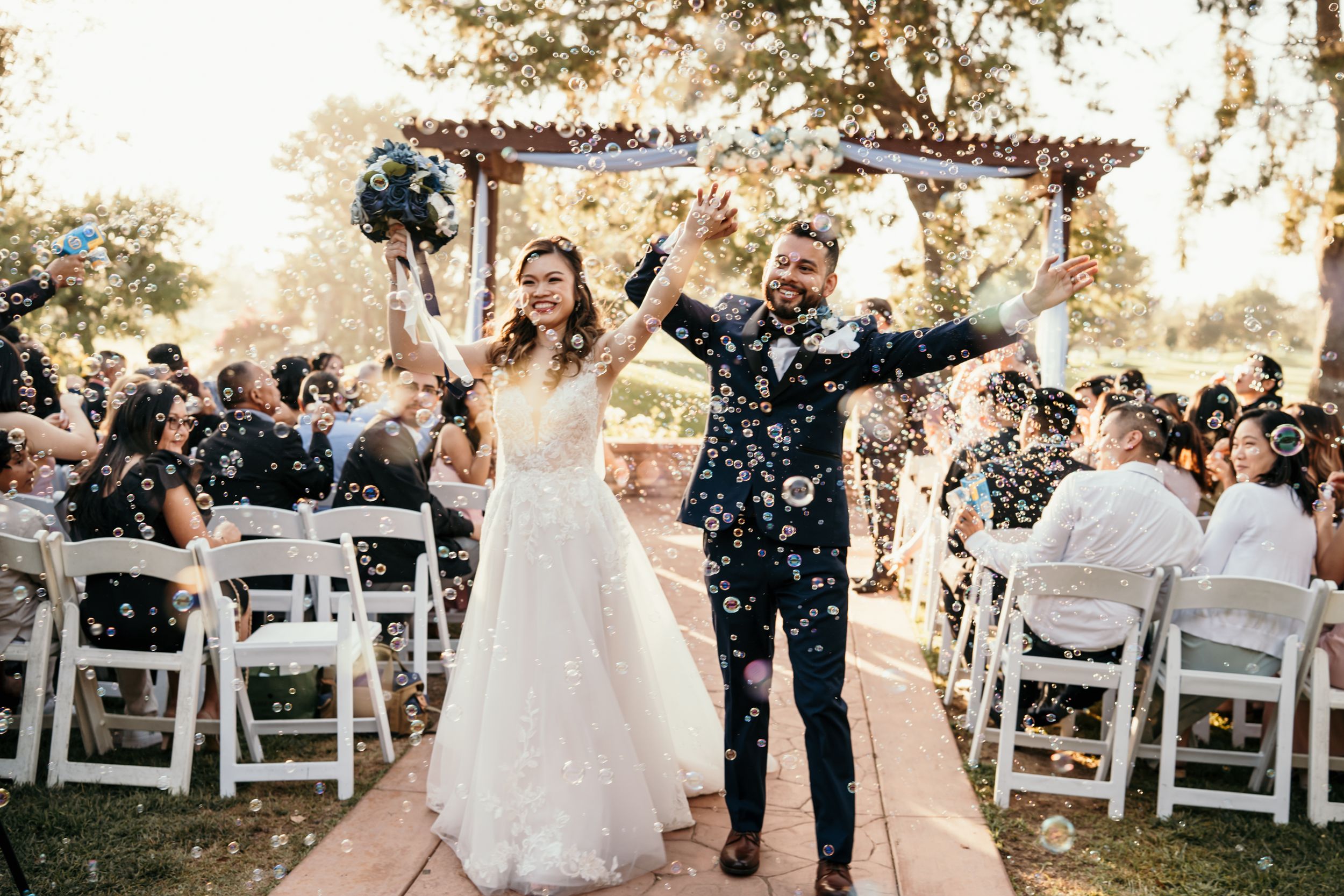 Couple celebrates under floral arch while guests throw confetti at outdoor wedding ceremony.