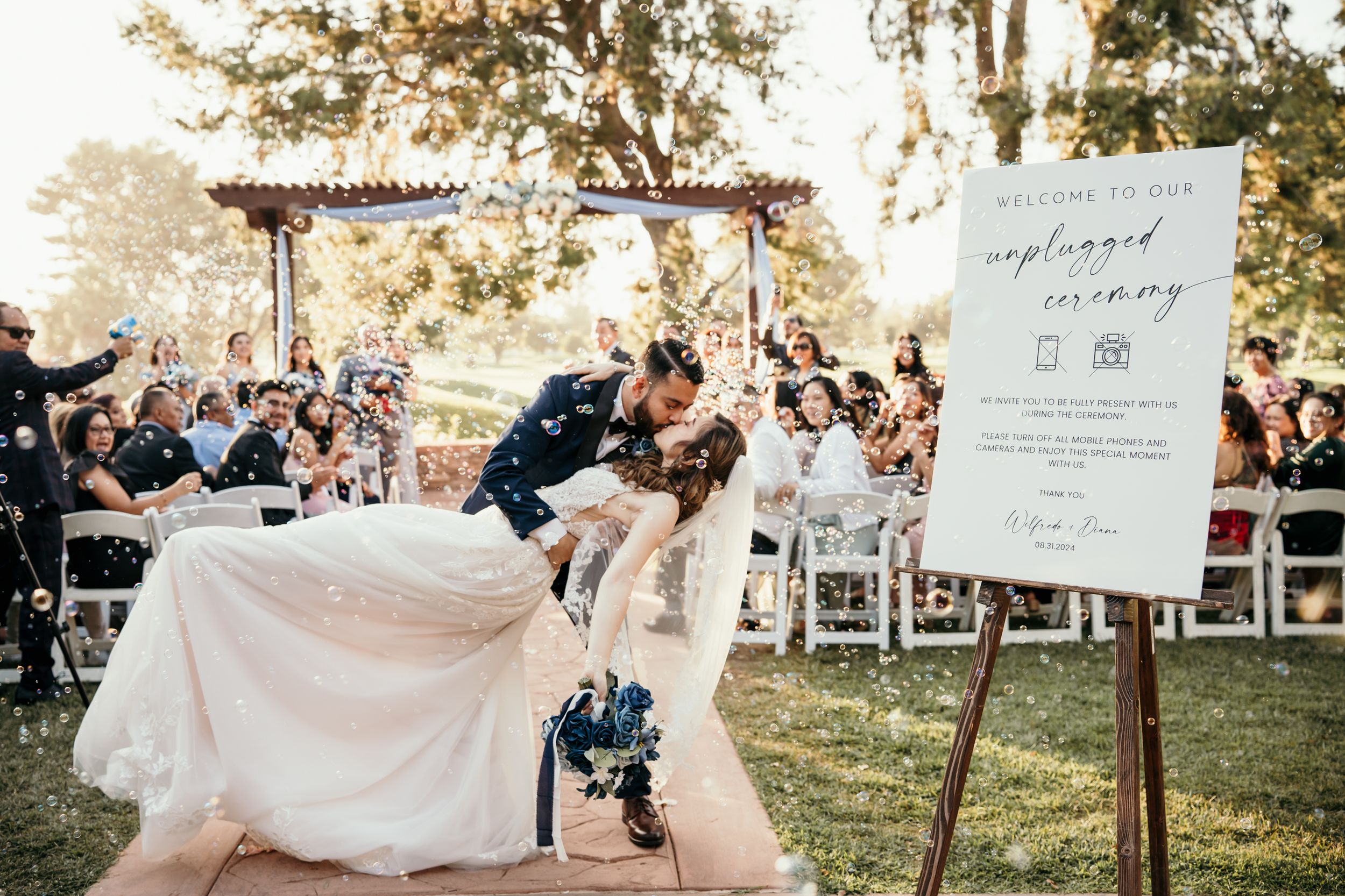 Wedding couple performs fun dip pose on aisle during outdoor ceremony.