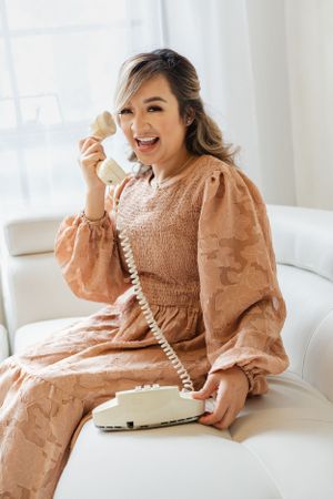 Woman in vintage peach dress talks on retro telephone while sitting on white couch.