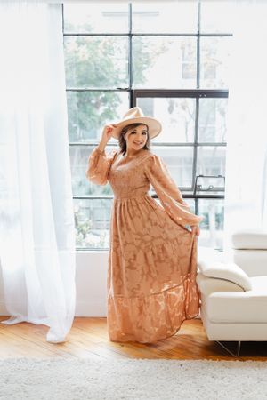 A feminine figure in a peach-colored maxi dress and wide brim hat poses elegantly by large windows in a bright room.