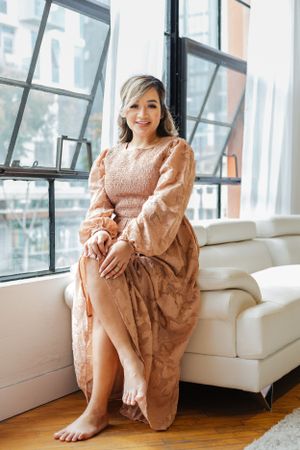 Person wearing a flowing peach dress relaxes barefoot on a white couch near sunny windows.