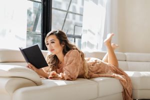 Person in peach dress reading book while lounging on white couch by window.
