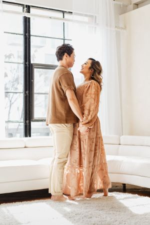 Couple in coordinating peach and brown outfits embrace near bright window.