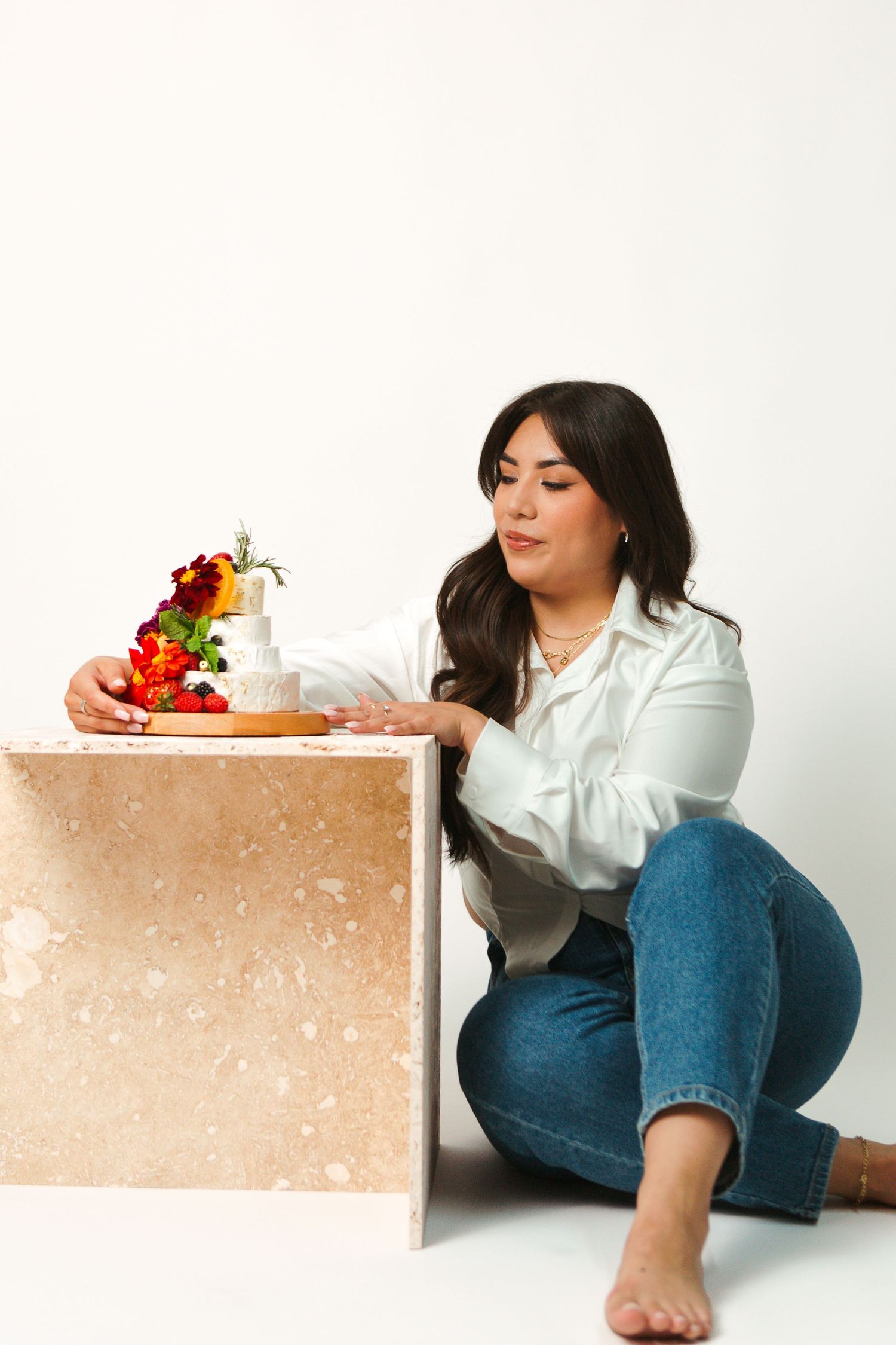 Woman in white blouse and jeans sitting with colorful flowers on wooden platform against white background.