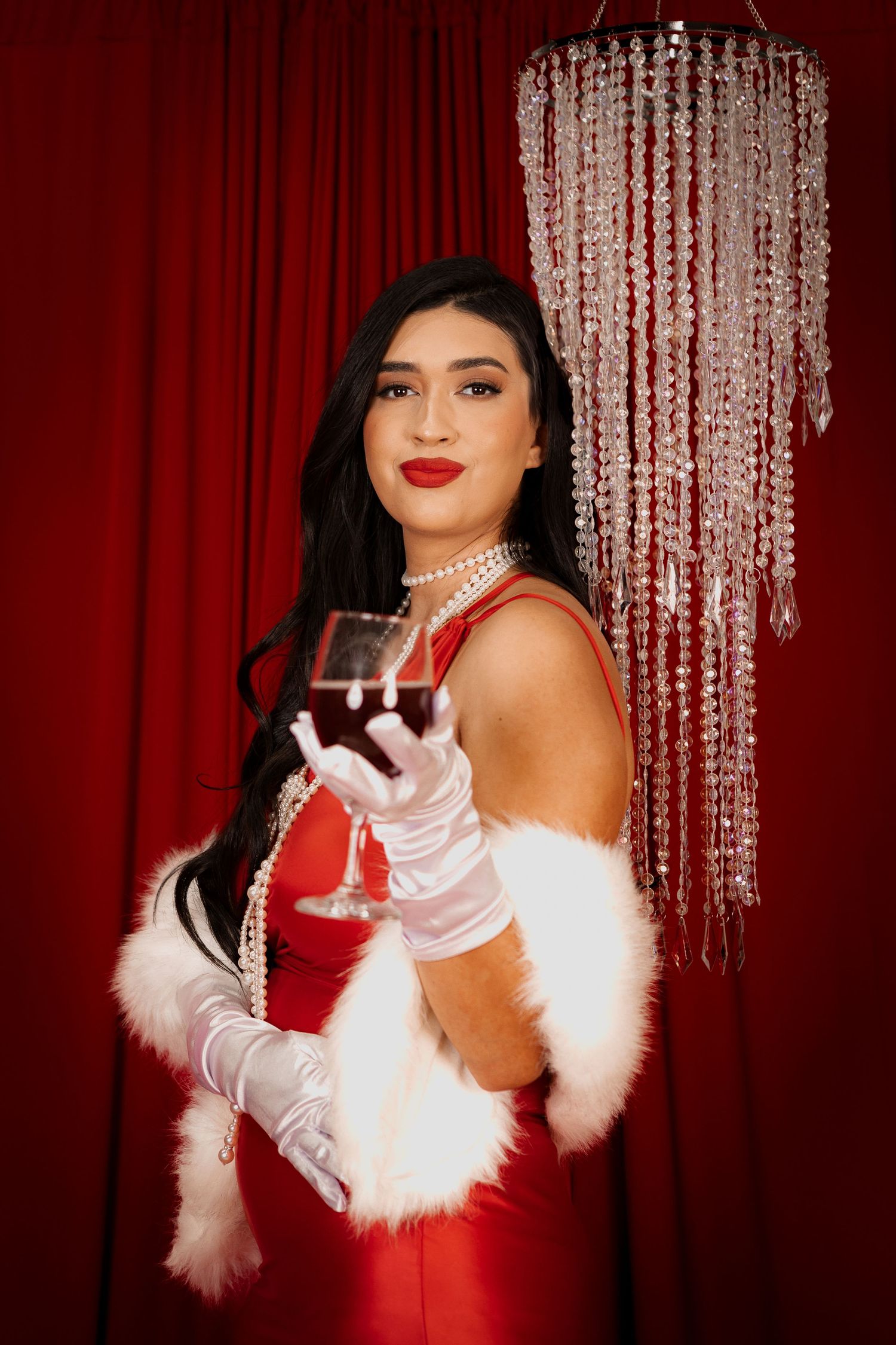 Elegant figure in red gown and white gloves holding wine glass against red curtain backdrop.