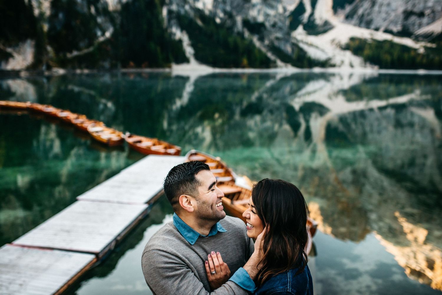 A couple laughing as he caresses her face with the lake, pier, boats and mountains behind them in Lago Di Braies in Italy.