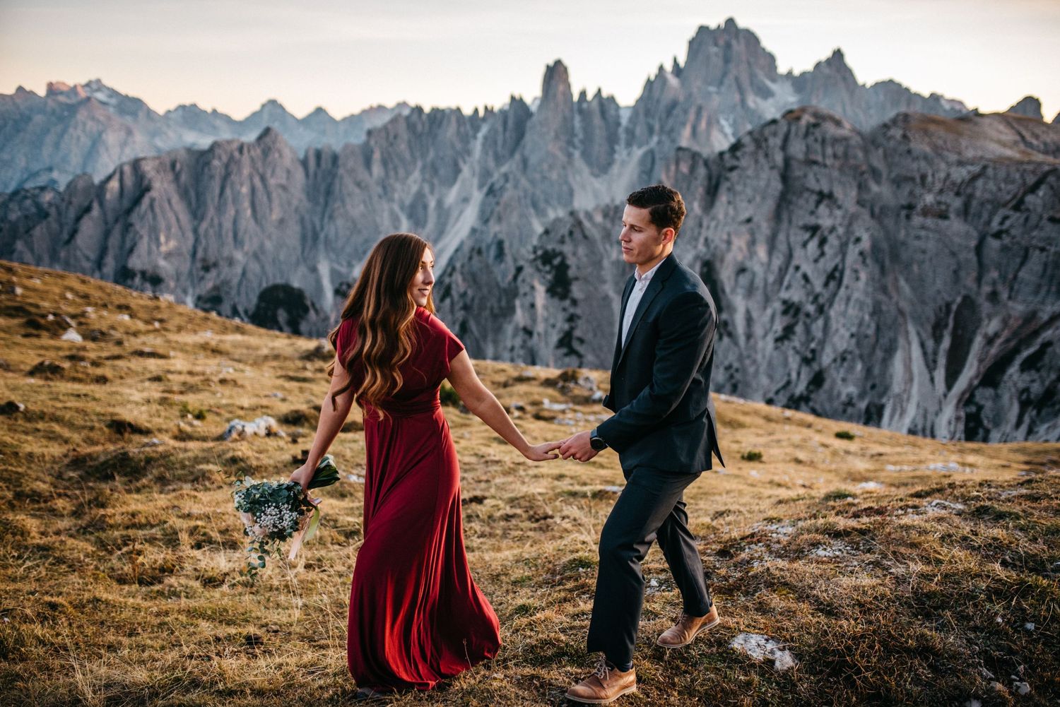 Wife is leading husband on a walk with the mountains behind them at Tre Cime, Italy.