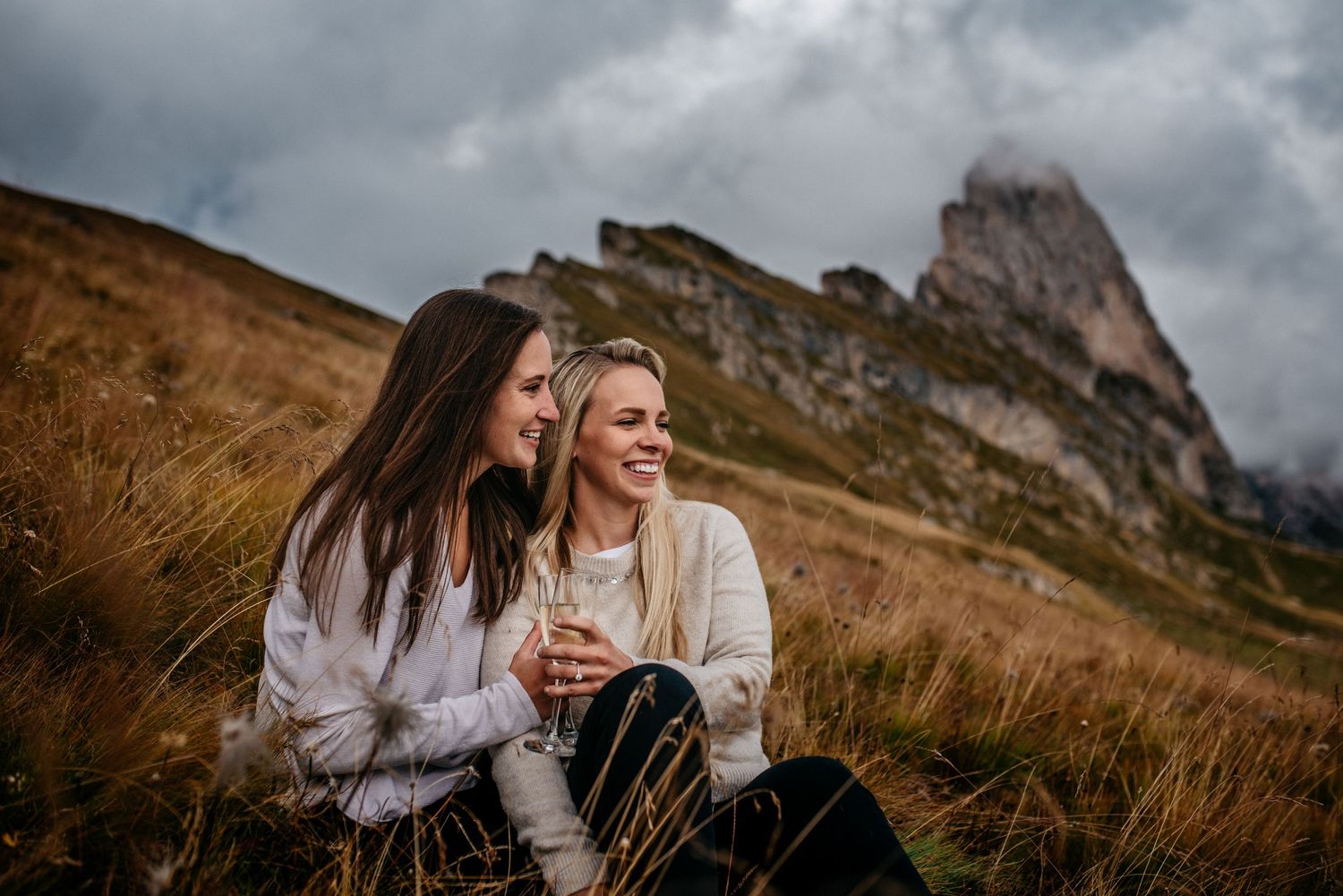 same sex couple sitting in the field in front of the mountain of Seceda sharing wine in Itay.