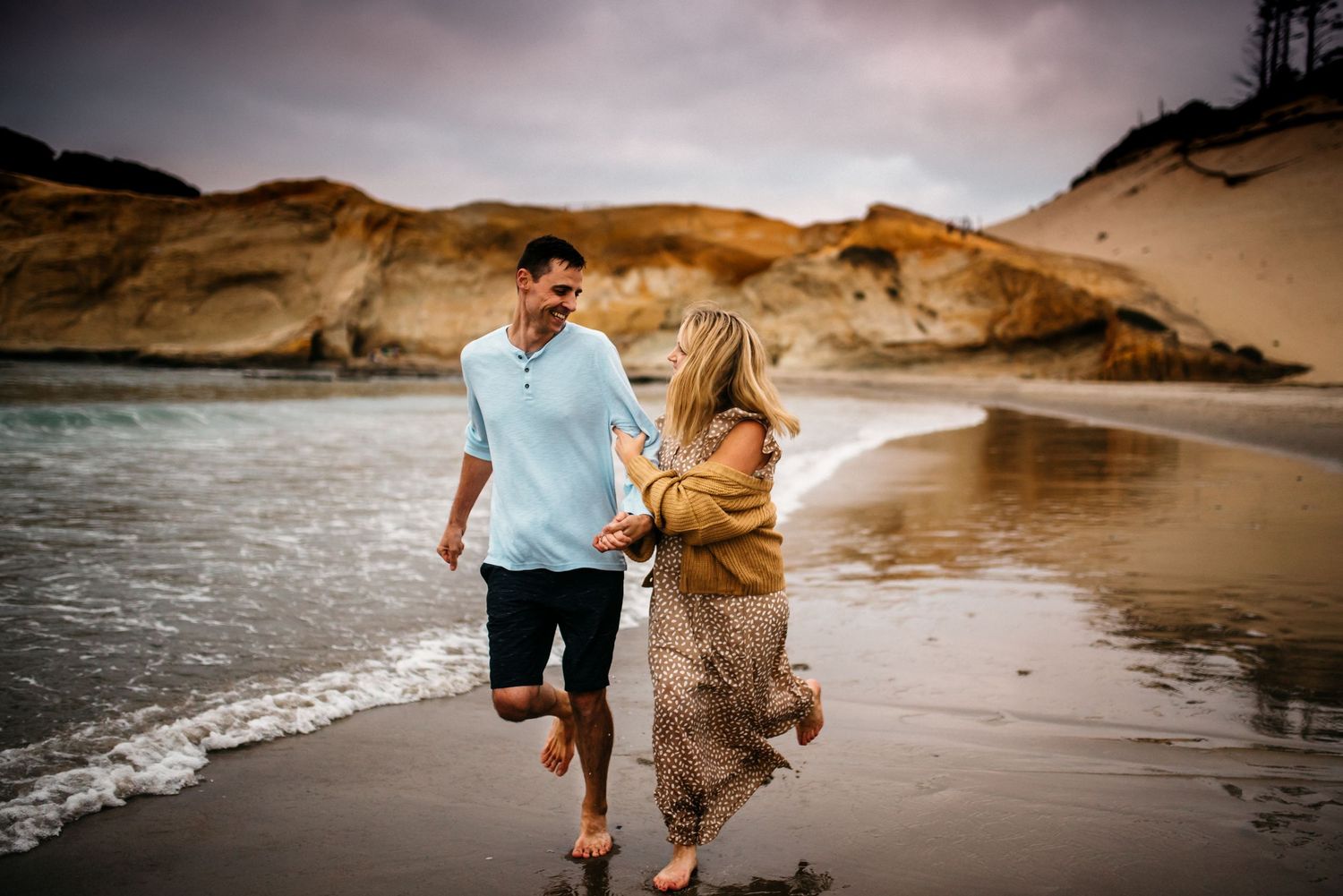Couple running among the waves with the rock formations behind them in Pacific City, Oregon.