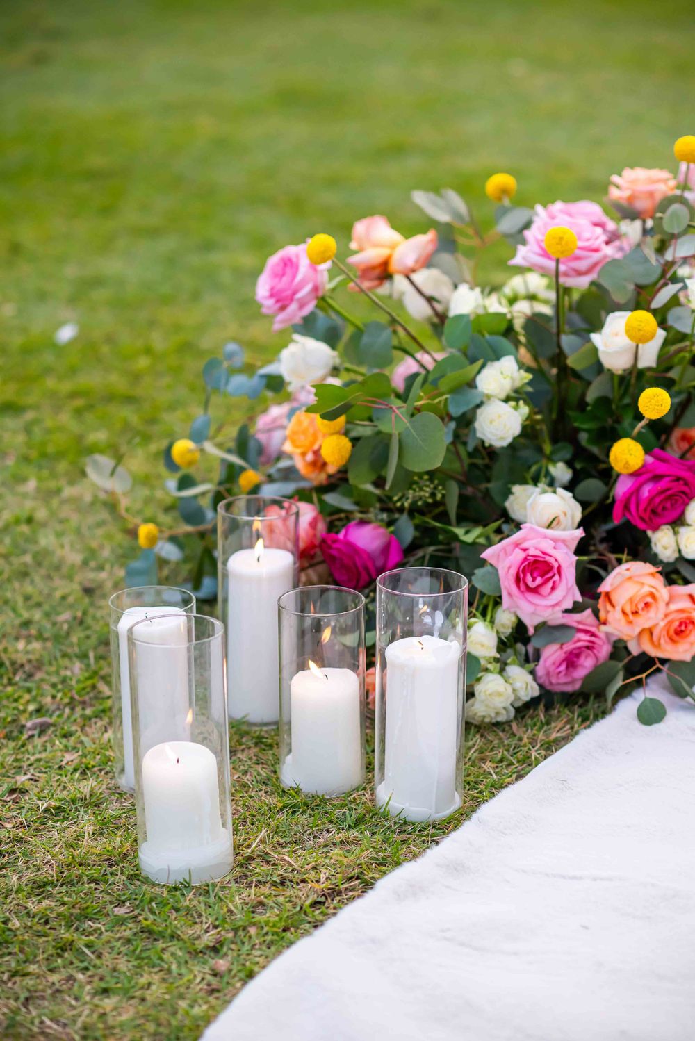 Elegant wedding decor with colorful roses and glowing candles in glass holders on grass.