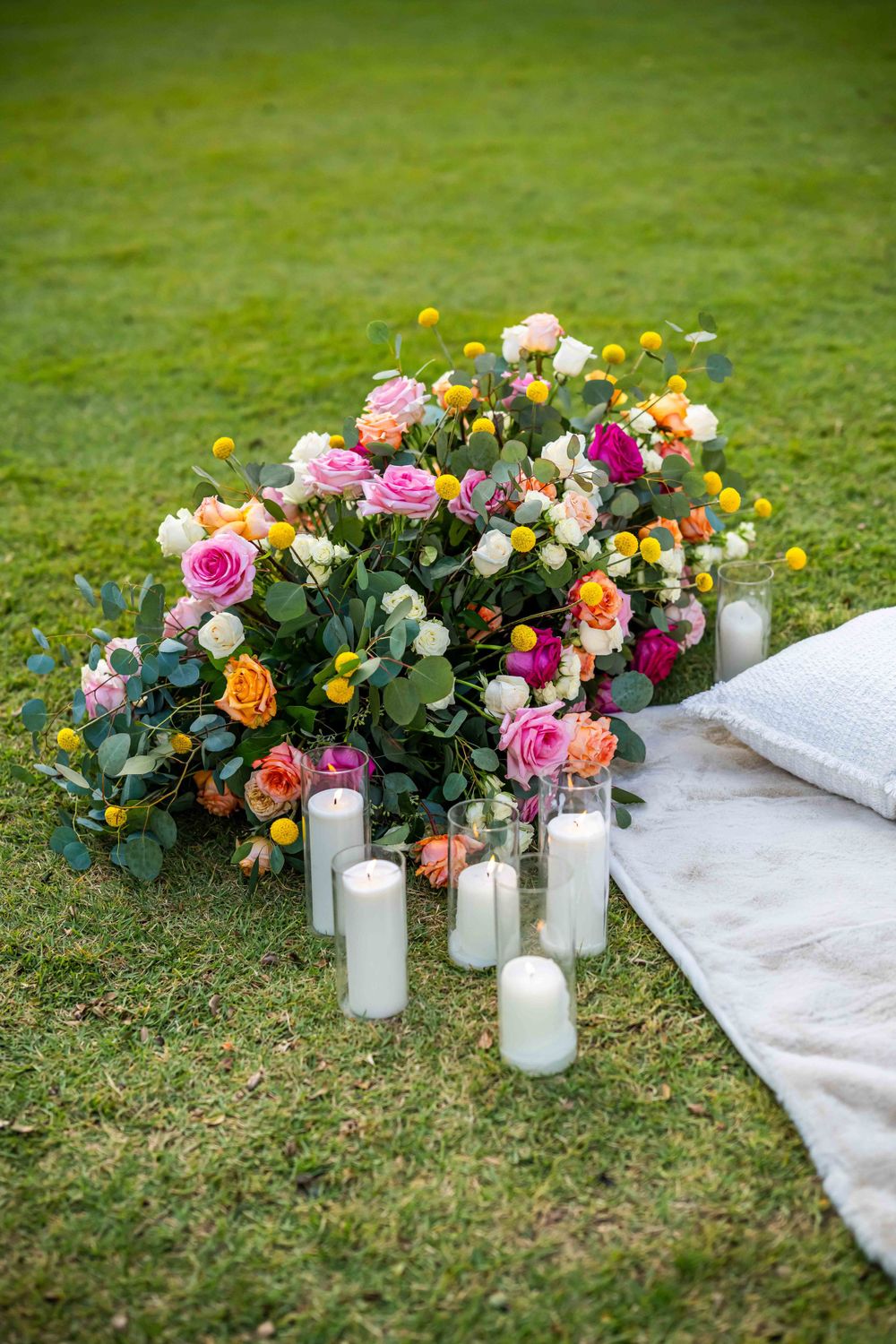 Colorful rose arrangement with white candles beside white cushions on green grass.