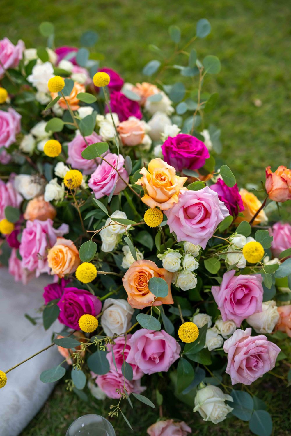 Vibrant floral arrangement featuring pink, orange, and yellow roses with eucalyptus greenery on a grass background.