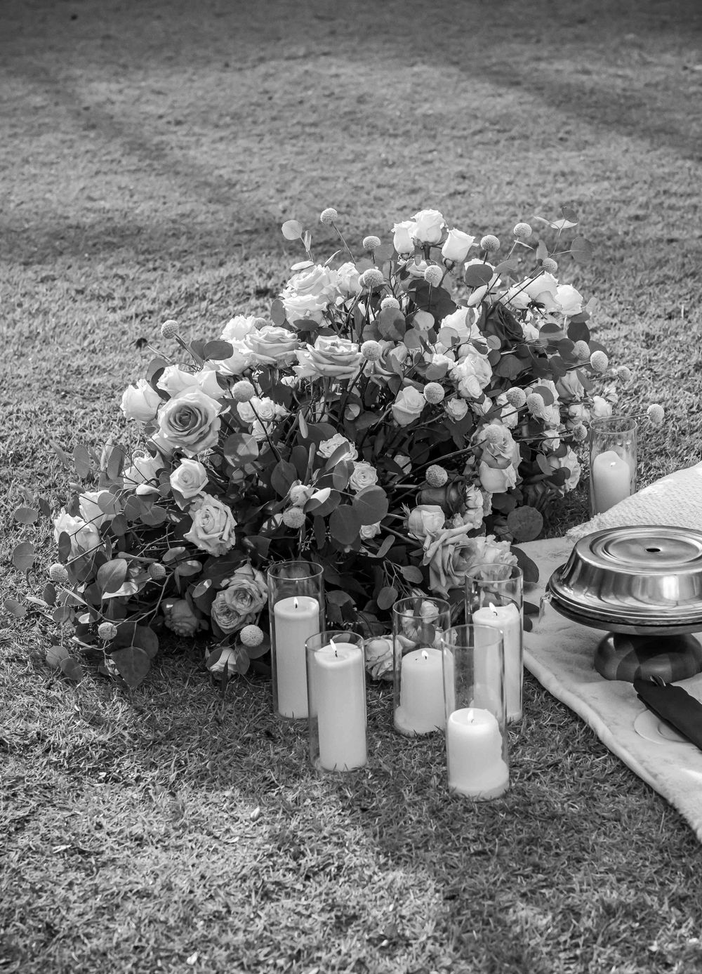 Black and white image of floral arrangement with pillar candles on grass setting.