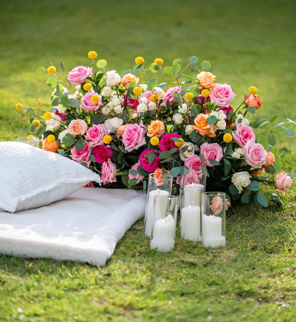 Pink and orange rose arrangement with yellow accent flowers and white candles on grass.