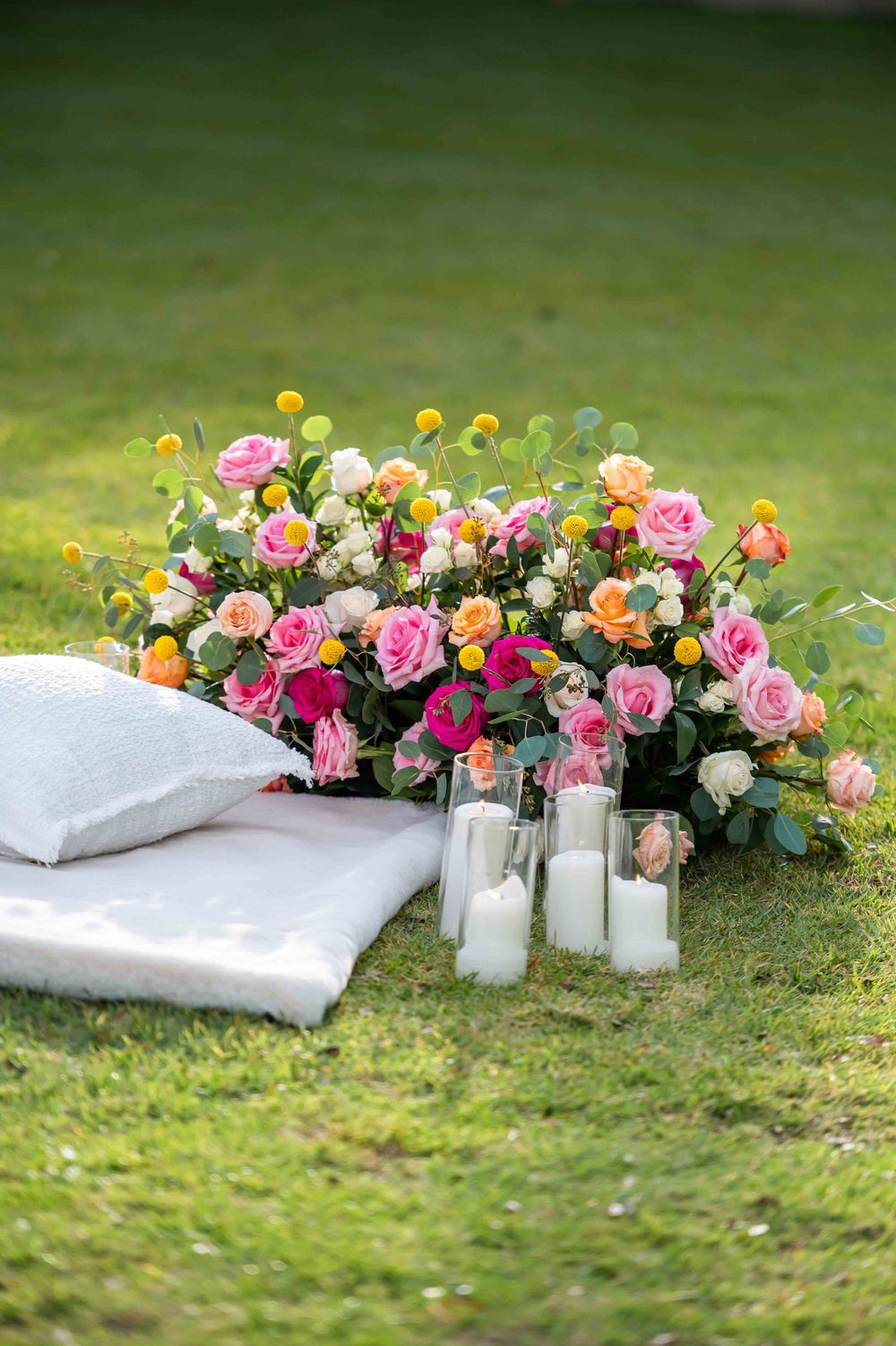 Romantic outdoor setting with pink rose arrangement, white pillows and glowing candles on grass.
