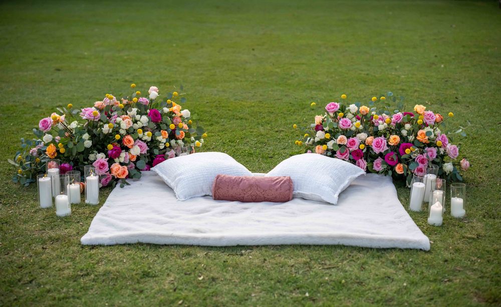 Romantic outdoor seating area with floral arrangements, pillows and candles on grass.