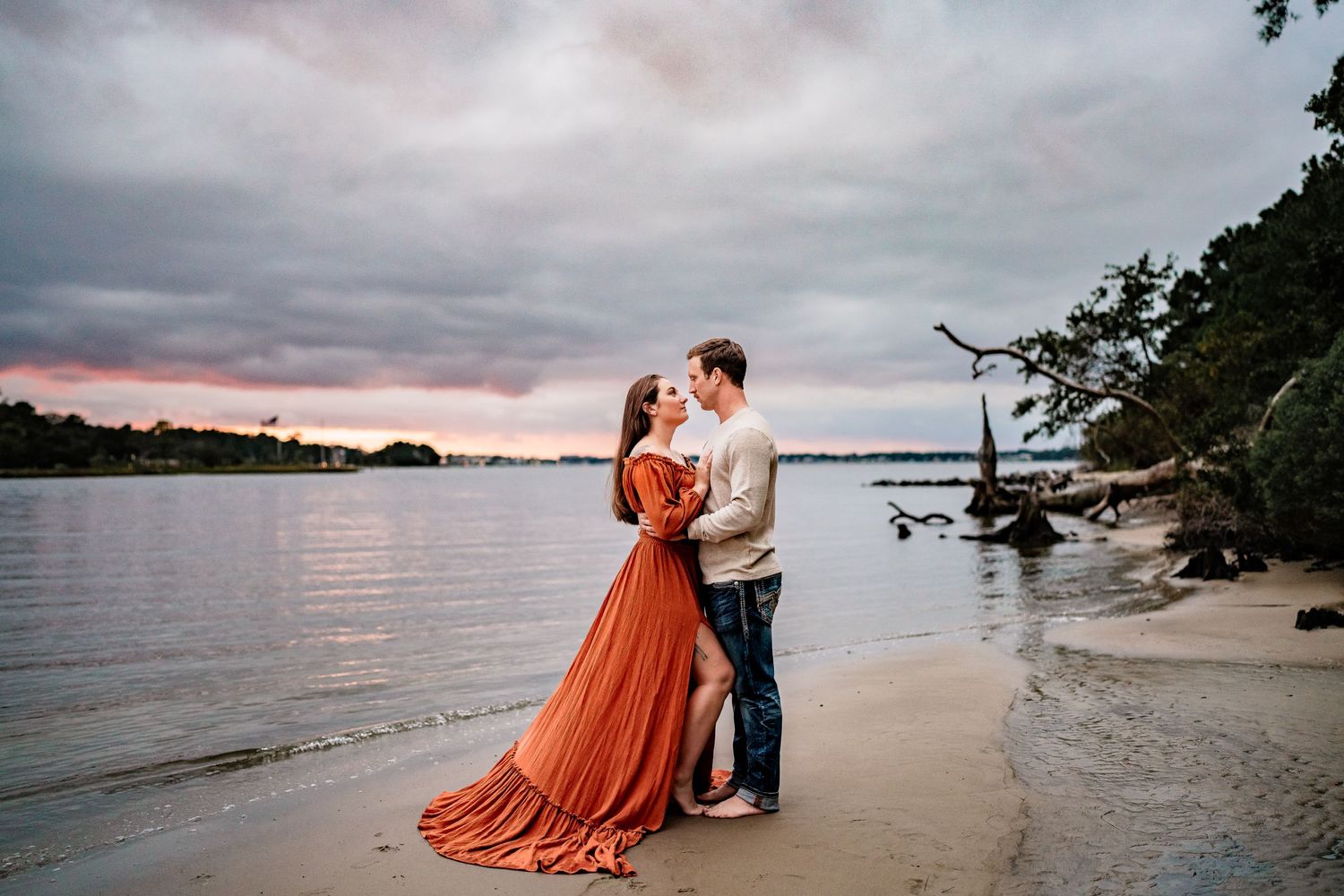 Couple in formal attire embracing on beach at sunset with dramatic cloudy sky.