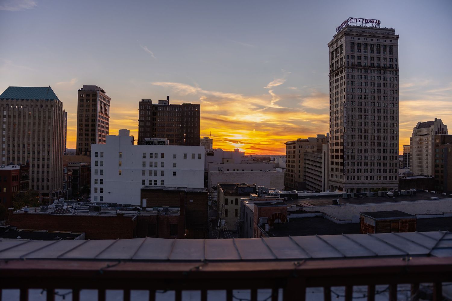 Panoramic cityscape view at sunset with tall buildings silhouetted against an orange and purple sky.