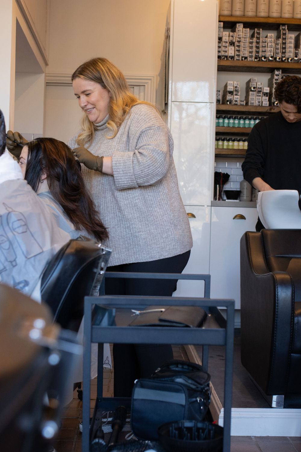 Professional hairstylist working in a modern salon with retail products displayed on shelves.
