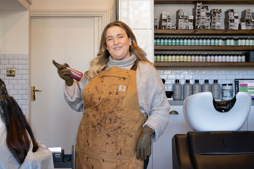 Stylist wearing brown apron holds tools while working in modern salon setting.