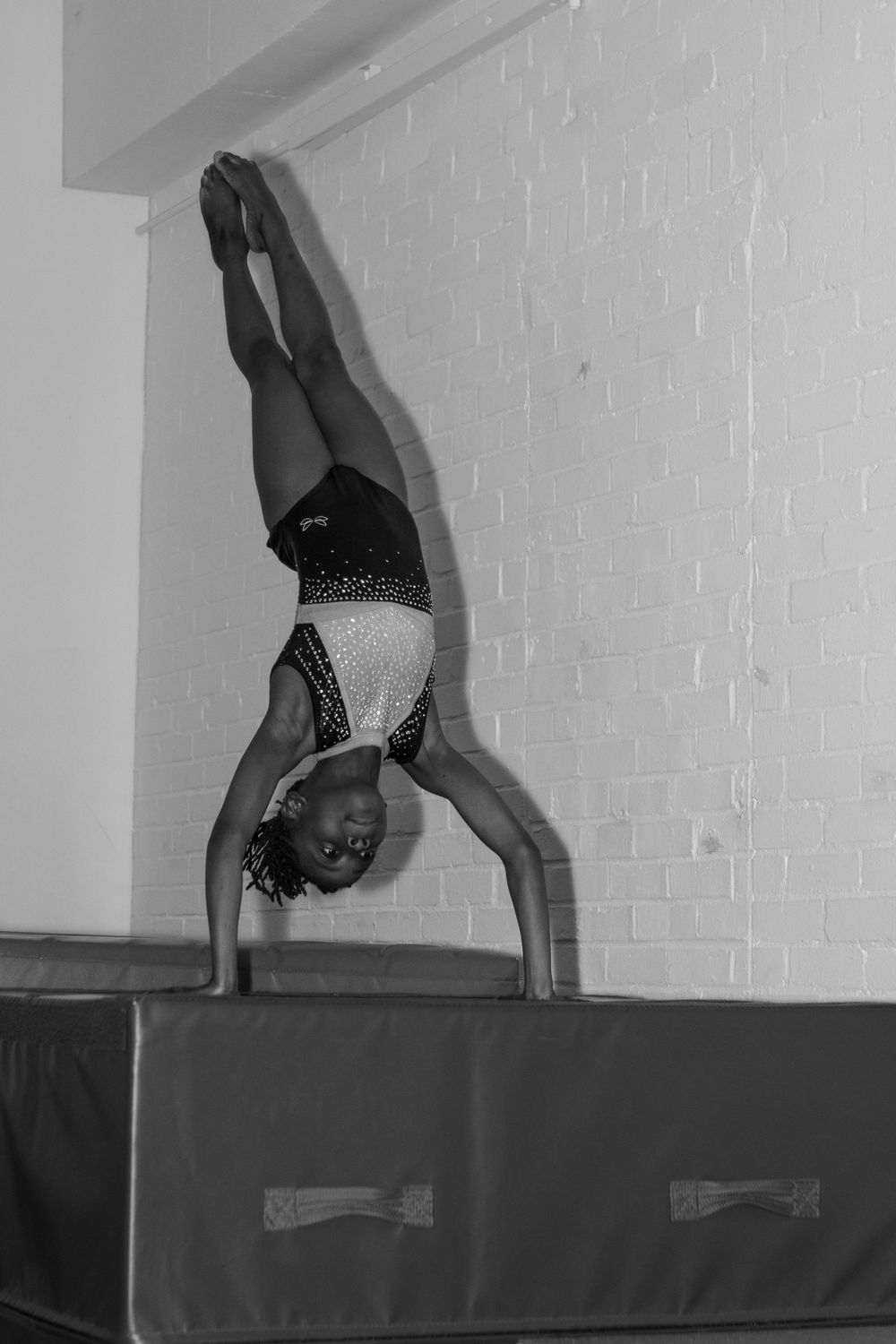 Gymnast performing handstands on a padded mat against a white brick wall.