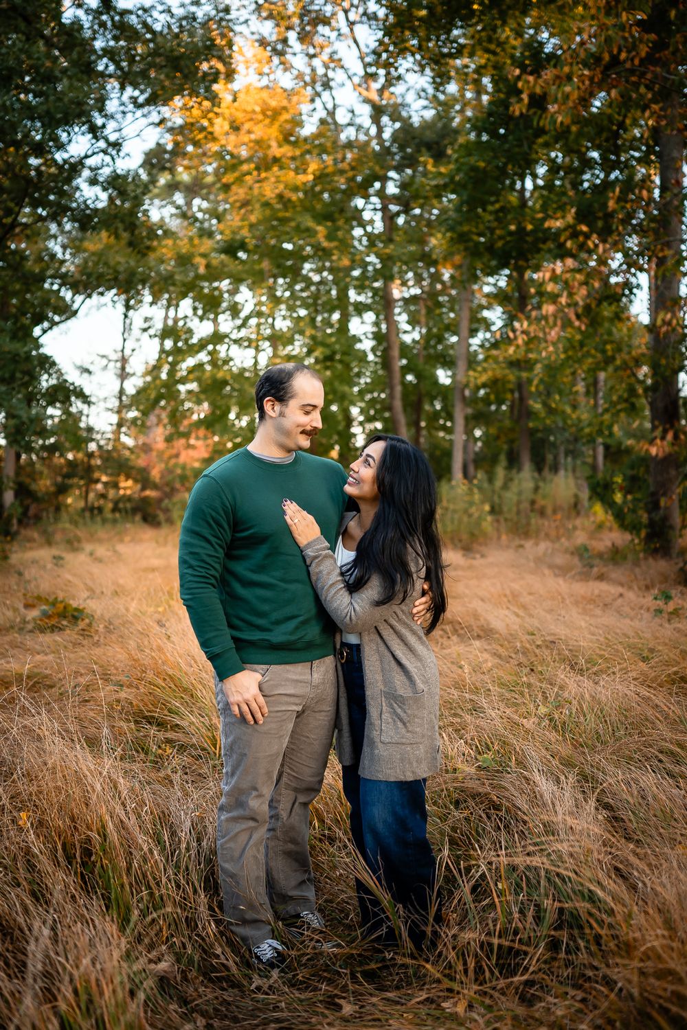 A couple poses romantically in a field of tall grass during golden hour with fall foliage behind them.