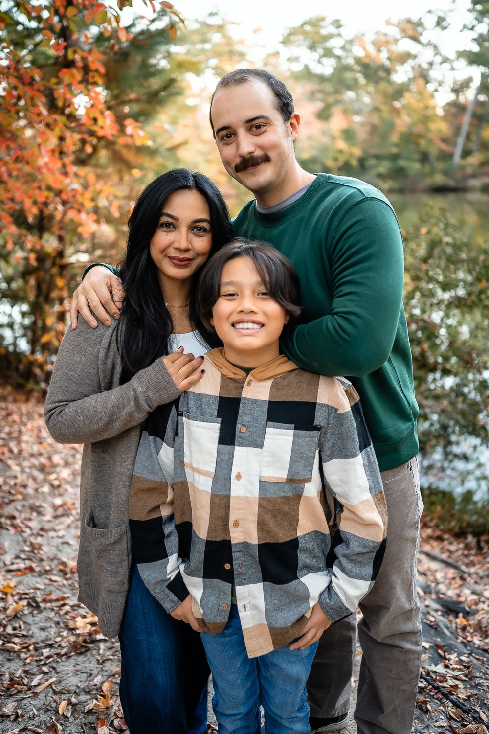 A family poses outdoors in fall foliage wearing casual clothing while standing near a body of water.