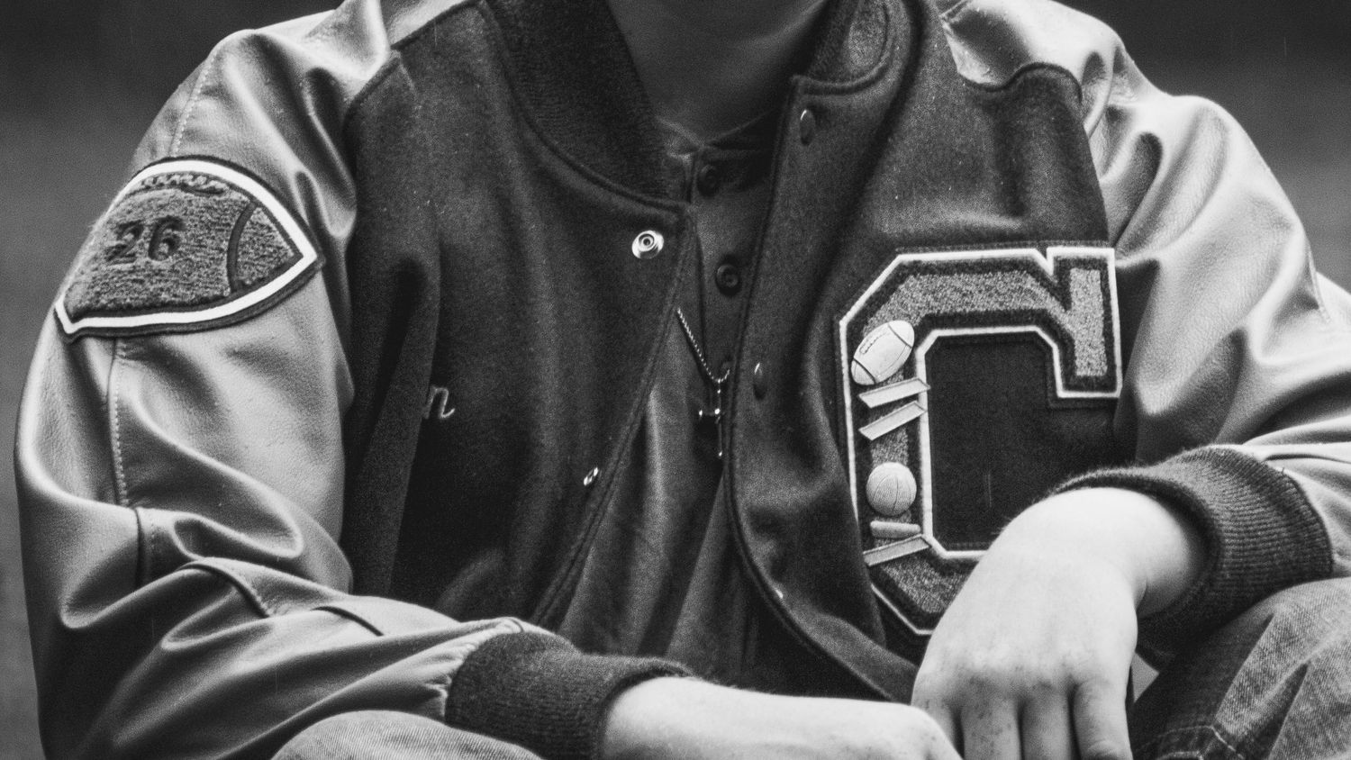 black and white image of young mans letterman jacket. a football patch is on one sleave and the Letter has 6 pins on it