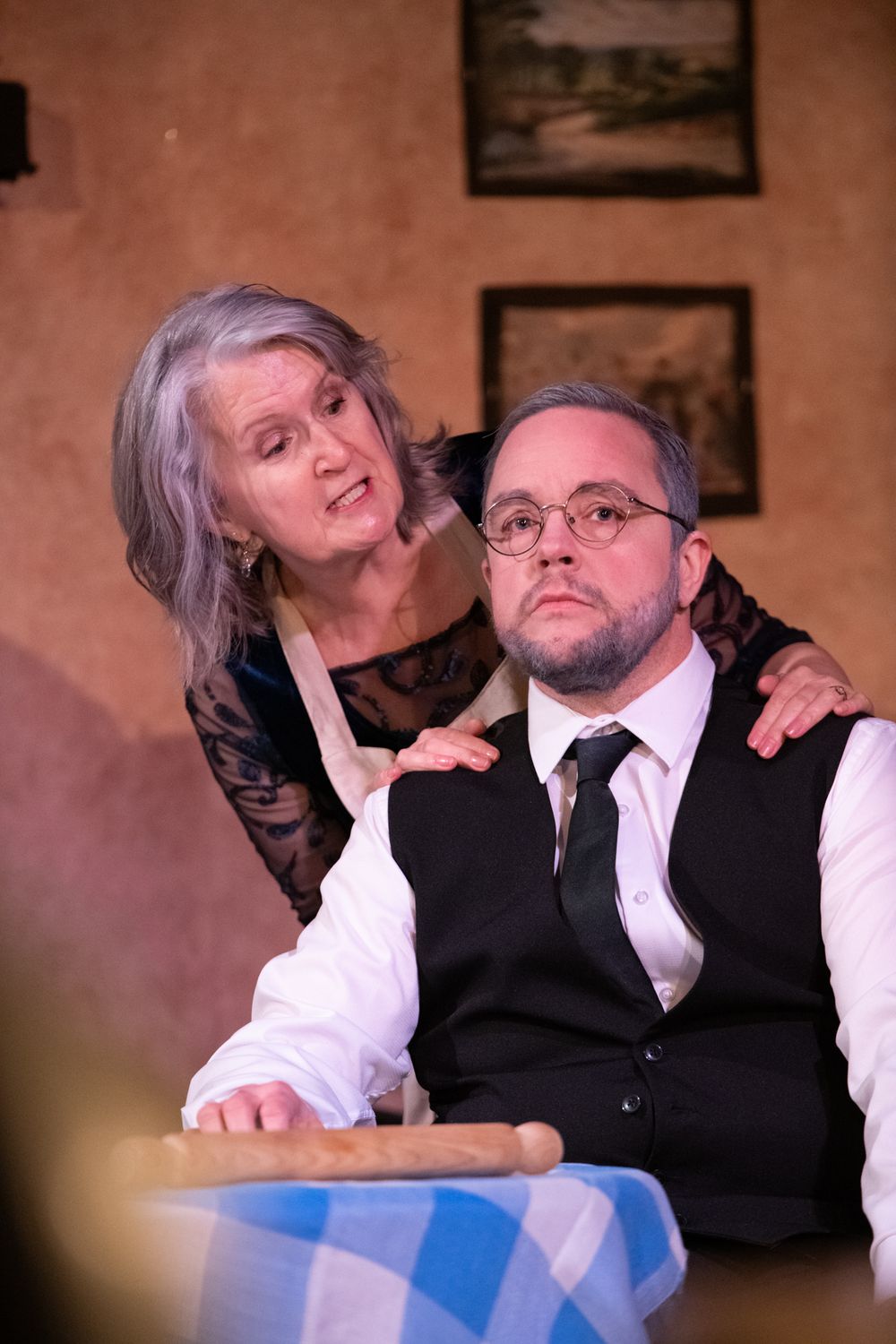 Two actors in a dramatic theater scene, woman in apron standing behind seated man in vest.
