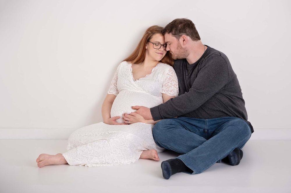 Pregnant young woman in white lacy dress sits with husband hugging eyes closed in studio Baltimore Ontario