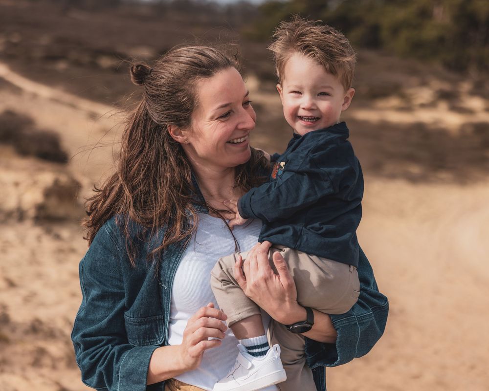 A smiling woman holds a laughing toddler boy outdoors on a sandy beach, both wearing casual clothing.
